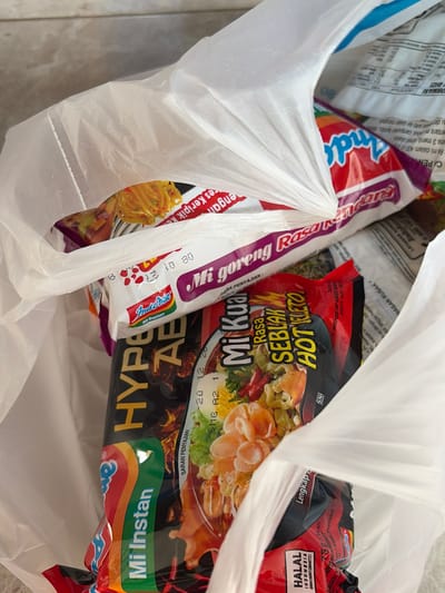 This image captures a close-up view of a white plastic shopping bag filled with instant noodle products. The bag is the primary object, and the focus is on the contents within. The scene appears to be indoors, likely a kitchen or countertop, as suggested by the neutral background.

Several brands of instant noodles are visible, including "Indomie Mi Goreng Rasa Seblak Hot Jeletot" and "Indomie Mi Instan Hype AB". The packaging is colorful, with prominent text in Indonesian such as "HALAL", "INDONESIA", "Mi Instan", and "Rasa Seblak Hot Jeletot". There are also nutritional and preparation instructions on some of the packaging. The "Indomie" logo, with its distinctive rainbow design, is clearly visible.

The lighting suggests daytime. There are no people visible in the image, and the focus is solely on the objects. The overall impression is a snapshot of groceries, specifically a collection of popular Indonesian instant noodle varieties. The location context of Denasri Kulon, Indonesia, aligns with the presence of Indonesian language text on the product packaging. The image is crisp and clear, allowing for detailed observation of the product labels.