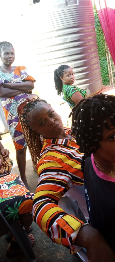 The image depicts an outdoor or open-air social gathering in Udi, Nigeria. In the background, a large, dark, corrugated cylindrical water storage tank is visible on the right, adjacent to a section of green foliage or artificial turf, partially obscured by vertical red fabric drapery. The ground is a light-colored, uneven surface.
Six individuals are present. On the left, a woman stands with her arms crossed, wearing a colorful short-sleeved dress with a geometric pattern in orange, purple, teal, and white. She has a white beaded necklace and small earrings, and her hair is dark and braided.
In the mid-ground, a woman is seated, facing slightly right but looking directly at the camera with a neutral expression. She wears a long-sleeved button-up shirt with horizontal stripes in yellow, orange, black, and white. Brown-tinted sunglasses rest on her light brown braided hair, and she has visible dangling earrings.
Behind and to the right of the central seated woman, another woman is seated, turned with her back mostly to the camera but looking over her right shoulder. She wears a green and black vertically striped top and has dark braided hair styled in a high ponytail, with hoop earrings visible.
In the foreground on the right, only the upper portion of a fourth individual's head and right shoulder are visible. This person wears a dark top with a pink collar trim, and their dark braided hair is adorned with numerous small, clear, spherical beads.
At the bottom left, the legs and a portion of a fifth individual are visible, seated, wearing a black dress with a prominent orange floral and green leaf pattern. Their feet, with painted toenails, are visible in dark sandals. A sixth individual's arm and hand are barely visible behind the woman in the striped shirt, suggesting further presence.