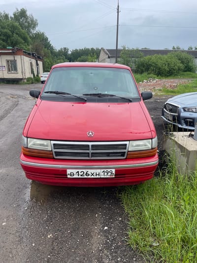 The image shows a red Dodge Caravan parked on an asphalt surface. The car's front is facing the viewer, and its license plate reads "B 126 XH 199 RUS". The background includes green foliage, a utility pole, and a partially visible building. The asphalt surface is wet, suggesting recent rain. Another car, possibly light blue, is partially visible parked behind the Dodge Caravan. Given the license plate's region code of 199, which corresponds to Moscow, it is likely that the location is in Moscow or the Moscow region, not specifically Khar'ino. The scene appears to be a roadside parking area or a residential street.