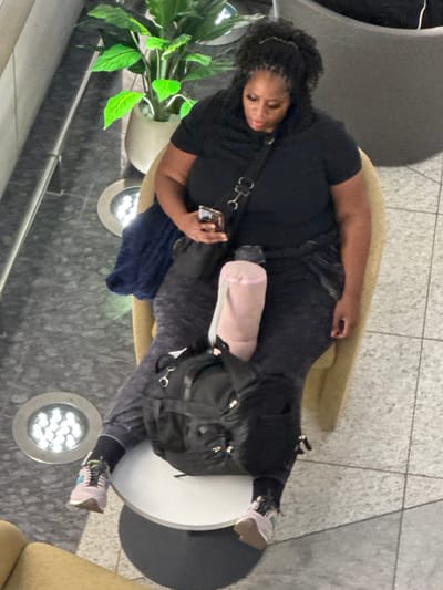 An adult female is depicted seated in a light tan armchair within an indoor facility in Al Matar, United Arab Emirates. She possesses dark, curly hair styled partially upward and wears a black short-sleeved top, dark grey patterned trousers, and light-colored athletic shoes with dark socks. Her attention is directed downward at a smartphone held in her right hand. A black crossbody bag is slung over her left shoulder. A folded dark blue garment is draped over the left armrest of the chair. A light pink, cylindrical object rests on her lap. A black backpack is positioned on a small, round table with a white top and a dark circular base directly in front of her. The floor is composed of light and dark grey tiled sections. Two circular, floor-mounted LED light fixtures are visible in the foreground to the left. A potted plant with green foliage in a light-colored planter is visible in the background to the left. Partial visibility of another similar armchair is noted in the lower left corner.