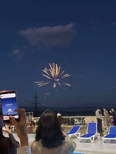 The image depicts an outdoor scene at dusk or night, with fireworks illuminating the dark blue sky. In the foreground, a person's hands are holding up a smartphone, which is capturing the fireworks display. The phone screen shows a digital representation of fireworks. Further back, a group of people are gathered on a deck or balcony, some standing and talking, others seated on lounge chairs. A prominent power pylon is visible against the sky, and distant lights suggest a settlement or city. The setting appears to be a gathering place with seating and railings, possibly a venue for events or entertainment. The overall atmosphere is celebratory, with the fireworks being the central focus of attention. The location is identified as being in Mukhatskaro, Georgia.