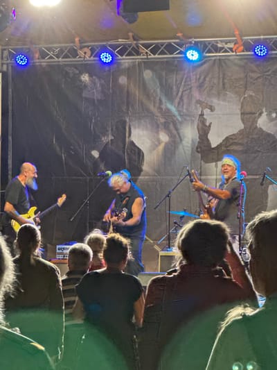 The image shows a live band performing on a stage. There are three male musicians visible, playing guitars and singing. The stage is set up outdoors, with a dark backdrop featuring silhouette graphics. Blue stage lights are illuminating the performers from above. In the foreground, the backs of several audience members are visible, watching the performance. A piece of audio equipment with "ORANGE" branding is visible on the stage. The overall scene suggests a concert or festival event. The location is identified as Tutrakan, Bulgaria, based on external information.