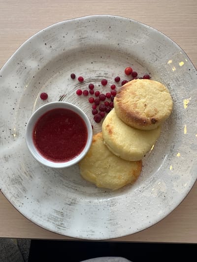 A close-up, overhead shot shows a white, speckled ceramic plate containing three round, golden-brown syrniki (cheese pancakes) stacked on top of each other. A small white ramekin filled with a vibrant red berry sauce sits to the left of the syrniki. Scattered around the plate are several small, red lingonberries. A dusting of white powder, likely powdered sugar, is visible on the syrniki and sprinkled across the plate. The plate rests on a light-colored wooden surface, suggesting a table. The background is softly blurred, but appears to be an indoor setting. There are no people visible in the image. The scene is likely from a cafe or restaurant in Slyudyanka, Russia, known for its natural beauty and local cuisine.