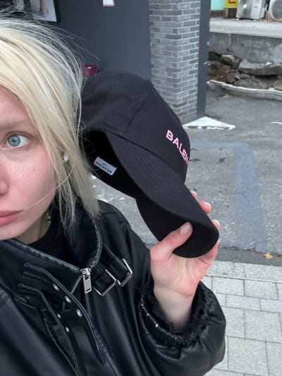 A blonde woman with blue eyes wearing a black leather jacket and a black Balenciaga baseball cap. She is holding the brim of the cap with her right hand. The street and some buildings are visible in the background.