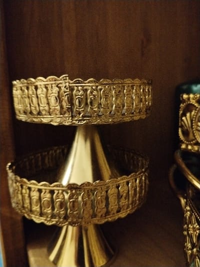 A two-tiered golden metal decorative stand features ornate detailing around the edges of each tier. The stand has a cone-shaped stem. Part of a second golden object, also ornate, is partially visible on the right side of the frame. The objects are placed against a solid brown surface that is out of focus.