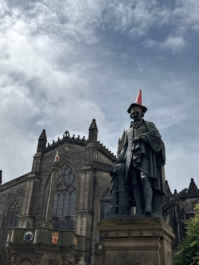 The photograph captures a bronze statue of Adam Smith, a prominent Scottish economist and philosopher, situated in the City of Edinburgh, United Kingdom. The statue is prominently positioned in the foreground, with a large Gothic-style building, likely a church or cathedral, serving as the backdrop. A striking detail is a bright orange traffic cone placed on top of the statue's head, suggesting a playful act of vandalism or a local tradition. The sky above is partly cloudy, with a mix of grey and white clouds against a blue sky. The statue is detailed, showing Smith in period attire, holding what appears to be a globe and a scroll. The building behind is made of stone and features architectural elements such as arched windows and spires. The overall lighting suggests it is daytime, possibly overcast.