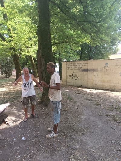 Two men, one white and one Black, are standing in a park in Casalmaggiore, Italy. The white man, on the left, is wearing a white tank top with "NYC" written on it and camouflage shorts. He is smiling and gesturing with his hands. The Black man, on the right, is wearing a light-colored t-shirt and denim shorts. He is holding his hands up as if in conversation. Behind them is a wall with graffiti. Trees and foliage are visible in the background, suggesting an outdoor, possibly urban, setting. The ground is covered with dirt, leaves, and some litter.