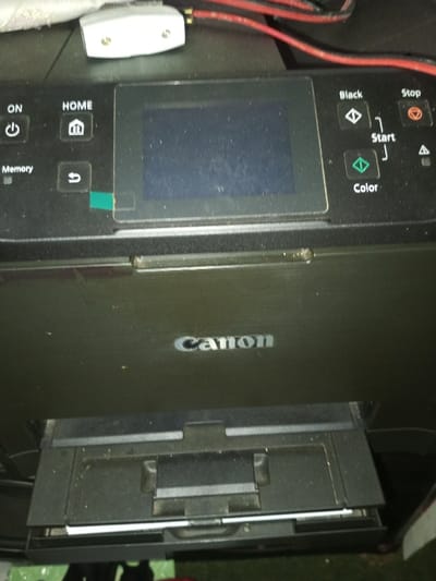 The image displays a black Canon printer. The control panel on top features an LCD screen, power, home, memory, back, black and color start, and stop buttons. A white power adapter rests near the top corner. A paper tray extends from the front of the printer. The Canon logo is prominently displayed in silver on the front panel above the paper tray. The printer appears dusty. Red cables are visible at the top.