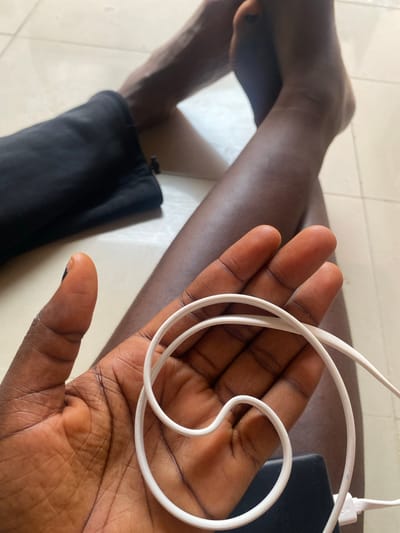 The image shows a person's legs and hand in Yola, Nigeria. The person is sitting on a tiled floor, with their legs crossed. Their left leg is partially covered by black fabric, likely shorts or pants. In their right hand, they are holding a white cable, possibly a charger or headphone cord. The cable is coiled and rests against their palm. The lighting suggests an indoor environment with natural light coming from the side.