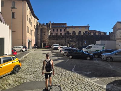 A person with a black backpack walks away from the camera on a cobblestone street lined with parked cars. The person is wearing a white tank top and black shorts. The scene is an outdoor urban setting with buildings on the left and a historic stone wall with battlements in the background. The weather appears sunny with a clear blue sky, suggesting it's daytime. The buildings vary in architectural style, with one being a light peach color and featuring arched doorways and shuttered windows. The stone wall is a prominent feature, likely part of an old fortress or castle. Several cars are parked on the street and in a lot, including a bright yellow hatchback on the left. Some visible text includes "Facility Forward. Il futuro è verde." and "honegger" on a white van. The overall impression is a warm, sunny day in a historic European town.