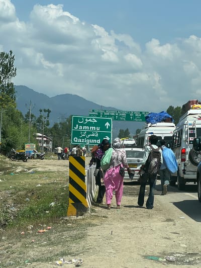 In Ujroo, India, a traffic scene unfolds on a dusty, roadside environment under a partly cloudy sky. A prominent green road sign indicates directions to "Jammu" and "Qazigund," with additional text in Urdu script. The sign also displays distances to "Banihal" (32 km), "Nashri Tunnel" (96 km), "Jammu" (191 km), and "Delhi" (805 km), along with "NH 44."
Several people are present. A group of individuals, some wearing traditional South Asian attire, are near the road sign. One woman in a pink salwar kameez carries a green bag, and another woman in a patterned headscarf and outfit walks beside her. A man with a backpack is also visible. Traffic is backed up, with several white vans and a car on the road. Some of the vans are laden with luggage on their roofs, covered with blue tarpaulin.
The roadside is unkempt, with visible trash scattered on the ground, including plastic wrappers and cups. A black and yellow striped marker post stands near the road. In the background, trees and mountains are present, with power lines and poles visible. Motorcycles are parked on the side of the road near a structure, and another sign for "Fastag Recharge Available Here" is visible. The overall scene suggests a busy transit point with an unmanaged roadside environment.