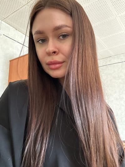 A young woman with long brown hair is centered in the frame. She is looking directly at the camera with a slight smile. She has light skin, grey eyes, and full lips. She is wearing a black garment. The background includes a wall with a textured, perforated surface and a light-colored tiled floor. Part of a wooden object is visible on the bottom left. There is no evidence in this image to indicate its location in Dubrova, Russia.