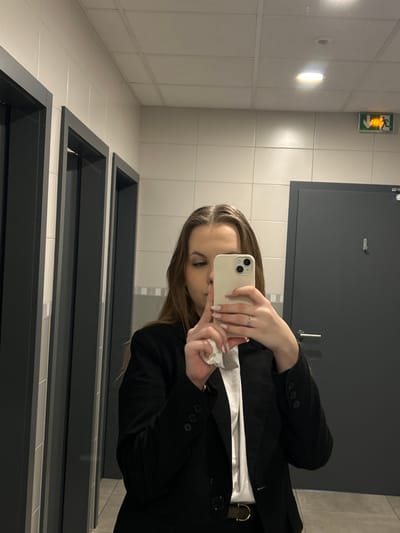 A mirror selfie is captured inside a public restroom in Reichstett, France. A female subject, with long light-brown hair, is visible from the chest up, reflected in the mirror. She is wearing a black blazer over a white shirt, with a dark belt featuring a metallic buckle visible at her waist. Her right hand holds a light-colored smartphone (appearing to be an iPhone model with dual rear cameras) horizontally, angled towards the mirror to take the photograph. A silver-colored ring is present on the fourth finger of her right hand, and her fingernails are painted with light-colored polish. Her left hand, partially visible, holds a crumpled piece of white tissue or paper below the phone.

The restroom features light beige or off-white square wall tiles, with a horizontal band of darker grey or blue tiles at mid-height. The ceiling is a suspended grid system with square panels, containing at least two recessed circular light fixtures, one of which is illuminated. An illuminated green emergency exit sign, depicting a running figure, is affixed to the wall in the upper right quadrant of the image. Multiple dark grey doors or partition panels are reflected in the background. One door on the right side of the image features a horizontal silver door handle and a vertical coat hook. Two additional dark grey doorframes or partitions are visible on the left side. The floor is not clearly discernible.