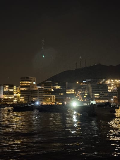 The image depicts a night scene in Ancón, Peru, with buildings illuminated along the coastline. The water reflects the city lights. The background shows a dark, mountainous landscape.