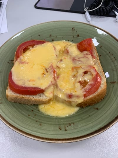 A close-up, eye-level shot shows a piece of toast topped with sliced tomatoes and melted cheese, presented on a green, speckled plate. The toast, a pale brown, forms the base for the vibrant red tomato slices, which are partially covered by a generous layer of melted, golden-yellow cheese. Some of the cheese has melted and dripped onto the plate. The plate is round with a textured, grooved surface and a dark brown rim. The background is blurred, suggesting an indoor setting, possibly a desk or table, with glimpses of office equipment like a computer mouse and cables. The overall lighting is bright and natural. There is no indication of the city of Urtaaul, Uzbekistan, or any people present in the image.