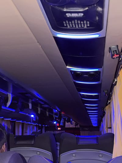 The image depicts the interior of a bus, likely a luxury coach, with a focus on the overhead compartments and seating. Blue LED lighting illuminates the ceiling and various vents and control panels. The seats appear to be upholstered in dark fabric, with headrests. The overall impression is one of a modern, well-equipped passenger vehicle. The mention of Bilaspur, India, suggests this is a specific location where such a bus might operate, though the image itself does not contain any explicit visual cues to confirm this location.