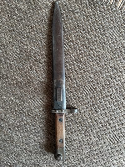 A bayonet with a worn, dark metal blade and a wooden handle rests vertically on a textured, woven brown surface. The blade exhibits signs of rust and age. The handle is secured with two dark rivets. A metal guard is present at the base of the blade. The background surface appears to be a mat or fabric with a repeating pattern of woven fibers. The lighting casts a subtle shadow to the right of the bayonet. There is no indication of the city of Osnabrück, Germany within the image.