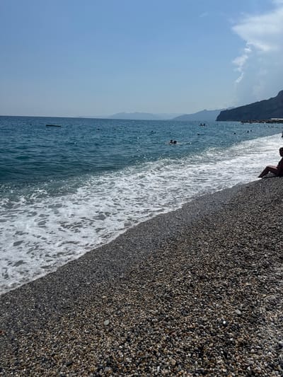 A sunny day at a pebble beach in Finale Ligure, Italy. The sea is a clear blue, with gentle waves lapping at the shore. Several people are swimming and enjoying the water, while others relax on the beach. In the distance, a rocky coastline rises up, dotted with greenery. The sky is a bright blue with a few wispy clouds. The scene is peaceful and idyllic, capturing the essence of a summer vacation.