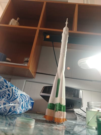A model rocket stands upright on a reflective surface, possibly a countertop. The rocket is primarily white with green and orange accents near its base, suggesting it is either a model of a rocket with those colors or is in the process of being painted. The rocket has a pointed tip and appears to be assembled from multiple segments.
Behind the rocket, a wooden shelf unit is visible. On the top shelf, a white ceramic object with a handle is present. The middle shelf has a box with the text "TES" partially visible, and beneath that, text that appears to be "Cosmop". A blue and white patterned bag or fabric is to the left of the rocket, partially obscuring a dark object.
In the background, a gray electronic device with a mesh front sits behind the rocket. To the right, a glass jar filled with a light green liquid and a stick is visible. The scene is indoors, illuminated by artificial light, as indicated by the bright glare in the upper right of the image. The overall impression is of a workspace or hobby area where the rocket is being constructed or displayed. There are no people visible. The time of day and weather are not discernible.