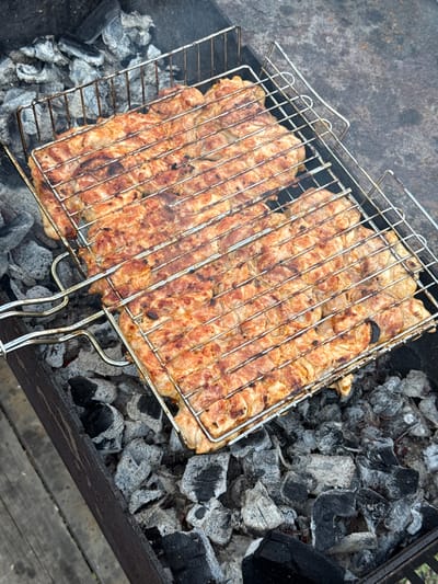 The image depicts a barbecue grill in what appears to be an outdoor setting in Кузнецово, Russia. The grill is filled with grey and black coals. Above the coals, a metal grill grate holds pieces of grilled meat, which appear to be cut into strips. The meat has a slightly browned color. The grill is positioned within a metal container, likely the barbecue itself, and part of a wooden surface is visible in the lower right corner, possibly indicating a table or platform. There are no people visible in the frame. The setting suggests a summer or warm-weather activity.