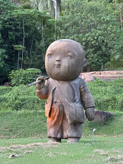 A bronze statue of a child with a large head, wearing a tunic and holding a stick, stands in a grassy area. The statue has a serene expression and appears to be illuminated from within by an orange glow. Behind the statue, lush green trees and foliage create a dense background. In the foreground, a small metal spotlight is planted in the grass. The scene is located in Baan Lipa Noi, Thailand.
