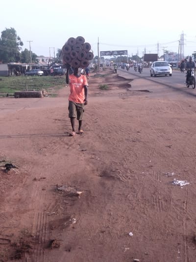 A man is walking on a dirt road in Kanshio, Nigeria (latitude: 7.717, longitude: 8.543). He is carrying a stack of round objects, possibly mats or rugs, on his head. The road is dusty and has tire tracks. There are several cars and motorbikes in the background, driving on a paved road. The scene is daytime and the weather is clear.