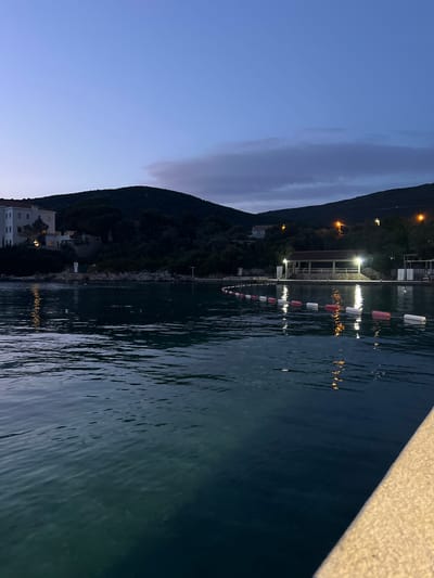 The image shows a nighttime view in Luštica, Montenegro. The body of water occupies a large portion of the scene, with light reflecting on its surface. A mountain stretches vertically along the left side of the image, featuring sparse structures and lights on its slope. A chain of white and red buoys spans horizontally across the water. A building or structure with lights is visible on the shoreline. A concrete edge is visible in the upper right corner.