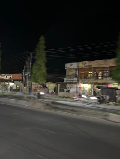 This is a blurry, low-light, handheld video clip taken at night, likely from a moving vehicle. The scene is a street in Bundar, Indonesia. The video is rotated 90 degrees clockwise, so the horizon appears vertical.
On the left side of the frame, there is a dark, open space, possibly a field or unlit area, with several power lines running vertically. To the right of the power lines, there is a row of buildings. These appear to be shops or businesses, with some windows illuminated from within. The architecture seems simple, with a mix of wooden and possibly brick facades. There are some signs and posters visible on the buildings, but they are too blurry to read clearly, except for one sign towards the bottom that appears to say "INTAS KUP". There are also trees or dense vegetation visible between the buildings and the power lines.
On the right side of the frame is a paved road, also blurry due to the motion of the vehicle. The road surface shows some wear and tear. The overall lighting is dim, with artificial lights from the buildings providing the main illumination. The blurriness suggests the video was captured quickly and without stabilization. There are no discernible people or specific activities taking place, other than passing by the scene. The time of day is clearly night, and the weather appears clear. The location cues are limited to the presence of shops, power lines, and a paved road, consistent with an urban or semi-urban street in Indonesia.
