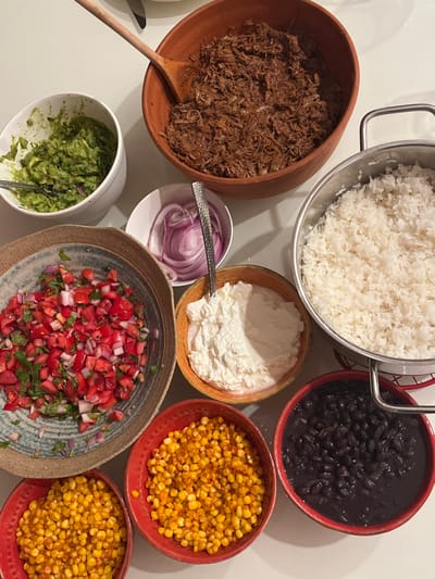 An overhead view displays a meticulously arranged spread of various food items on a light-colored, possibly white, tabletop. No individuals are present.
The following food items are presented in distinct containers:
* A large terracotta bowl contains a generous portion of shredded brown meat, with a wooden serving spoon resting within it.
* A silver pot with a metal handle holds a substantial quantity of cooked white rice.
* A white ceramic bowl contains a green, mashed mixture, consistent with guacamole, with a silver spoon partially submerged.
* A smaller white bowl holds thinly sliced red onions, accompanied by a silver spoon.
* A textured ceramic bowl, with a blue-grey rim, contains a fresh salsa-like mixture of finely diced red tomatoes and chopped green herbs.
* An orange-hued bowl contains a white, creamy substance, likely a dairy product such as sour cream, with a silver spoon.
* Two red, textured bowls are filled with bright yellow corn kernels.
* A final red, textured bowl contains black beans in a dark liquid.
The arrangement suggests a self-service meal setup. The specific location in the city of Merom HaGalil Regional Council, Israel, cannot be ascertained from the visual evidence provided.