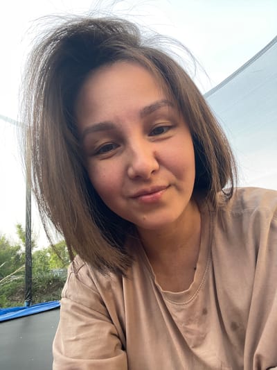The image features a close-up, self-portrait of a young adult female in an outdoor setting located in Verkh-Katunskoye, Russia. The woman, with a fair complexion, dark eyes, and medium brown, shoulder-length hair that appears slightly disheveled at the crown, is facing the camera with a subtle, closed-mouth smile. She is wearing a light beige short-sleeved t-shirt, which shows several wrinkles and two prominent dark, circular stains on the front left chest area. The background elements indicate a domestic backyard environment. A black mesh trampoline safety enclosure net is visible, curving across the upper right portion of the image and vertically along the left edge. Beyond the trampoline netting on the left, green foliage from trees or bushes is discernible against a bright, hazy sky. A blue tarp is partially visible on the ground to the left, beneath or adjacent to the trampoline. The lighting is natural daylight.