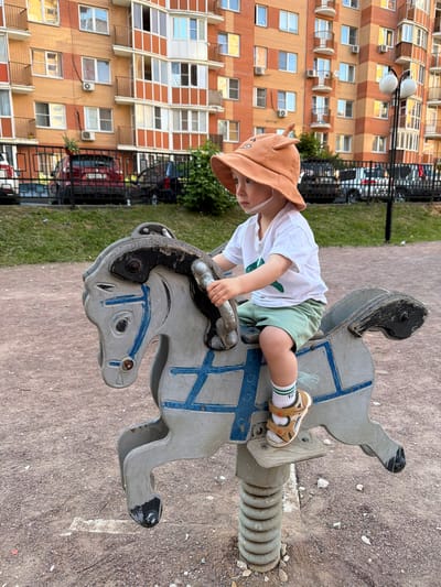 A young child, approximately a toddler, is depicted in a full-length portrait, seated on a grey, weathered wooden spring rider shaped like a horse. The child wears an orange wide-brimmed sun hat with cartoonish ears and a chin strap, a white short-sleeved t-shirt with a green graphic, light green shorts, white socks with green stripes, and tan sandals. The child's hands grip the handlebars of the spring rider, and their gaze is directed towards the left with a neutral expression. The horse-shaped rider features faded blue painted accents, including stylized crosses and facial details. It is mounted on a visible spiral coil spring, anchored into the ground.
The ground surface is composed of light brown, fine gravel or dirt with scattered small stones. In the mid-ground, a black metal fence with horizontal bars separates the immediate play area from a paved surface. Behind the fence, a small strip of green grass is visible, followed by parked vehicles, including the rear of a red SUV with a visible license plate and other partially obscured cars. A multi-branched street light with two white spherical lamps stands to the right of the fence.
In the background, two large, multi-story residential buildings dominate the scene. Their facades are characterized by alternating sections of orange-brown brick-like material and lighter tan or cream panels. Numerous windows with white frames and various balconies, some enclosed with glass and others featuring metal railings, are visible. Multiple external air conditioning units are mounted below windows on the building facades. The scene is illuminated by natural light, casting warm tones on the buildings. The setting is a playground within a residential area in the city of Vniissok, Russia.