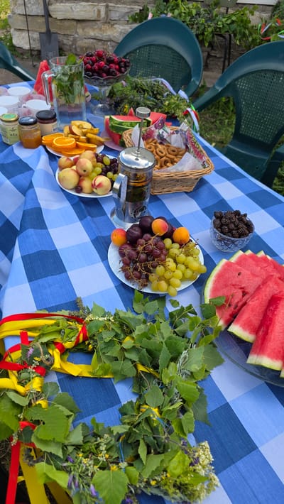 This image depicts an outdoor table setting in Kupoyarovo, Russia, laid with a blue and white checkered tablecloth. The table is laden with a variety of fruits, including sliced watermelon, peaches, plums, and bunches of green and red grapes. A glass pitcher filled with water and mint is also present, along with a glass bowl of cherries, a basket of small ring-shaped bread products, and a glass bowl filled with dark berries, possibly mulberries. A French press filled with tea or coffee sits on the table. In the foreground, a wreath made of greenery and adorned with red and yellow ribbons is laid out. The background shows a stone wall, green foliage, and two green plastic chairs. The overall scene suggests a gathering or picnic. No people are visible in the image.