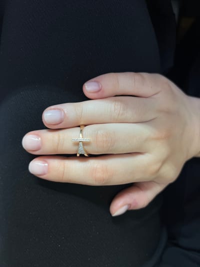 The media file shows a close-up of a person's left hand, adorned with a delicate gold ring. The ring features a minimalist design with a straight bar studded with small diamonds on one side, and a triangular cluster of diamonds on the other, connected by a slender gold band. The hand is resting on a dark, textured fabric, possibly clothing. The person's fingernails are neatly manicured with a subtle, sheer polish, reflecting a soft light. The background is dark and out of focus, drawing all attention to the hand and the ring. The lighting suggests an indoor setting, with soft illumination that highlights the details of the jewelry and the skin. There is no visible text in the image. The overall impression is one of understated elegance and personal adornment.