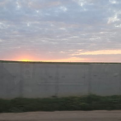 This image, taken in Ben Guerir, Morocco, depicts an outdoor scene at either sunrise or sunset. The upper half of the frame shows a sky with scattered altocumulus clouds, transitioning from light blue-grey at the top to soft pink and orange hues near the horizon. A bright yellow-orange glow, indicative of the sun, is visible just above the horizon on the left. The mid-ground features a long, light-grey concrete barrier extending horizontally across the frame, with faint reflections or marks on its surface. A dark, circular shape is visible on the left section of the barrier. Above the barrier, a flat, dark horizon line separates the sky from a faint strip of ground. Two thin, dark vertical structures, resembling utility poles or towers, are faintly visible on the horizon, one to the right and another towards the left. The immediate foreground shows a blurred strip of dark green vegetation, suggestive of grass, below the concrete barrier. Below the grass, a blurred, dark reddish-brown band represents a road surface or dirt track. No individuals or active subjects are visible in the scene.