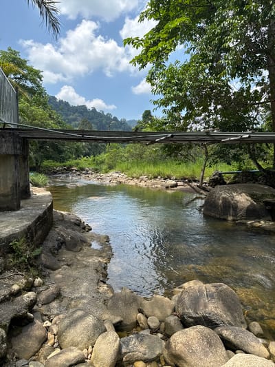 The image depicts a flowing river in a natural setting, likely taken in Tambon Song Phraek, Thailand. The river is bordered by rocks and vegetation. There is a man-made structure, possibly a bridge or support beam, spanning across the river. The sky is visible with scattered clouds. A mountain range is present in the background.