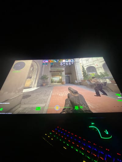 A photo of a monitor screen displaying a first-person shooter game, possibly Counter-Strike: Global Offensive, is captured from an oblique angle. The game scene depicts a street in what appears to be a European-style urban environment with buildings, trees in pots, and a brick-paved street. Two figures are visible in the game: one prone on a bench, and the player character holding a Glock-18 pistol. The player has $2100, 20 bullets, and 31 in reserve. The game interface shows the location as "MERCATO" and the player's health is at 100. The screen also displays "База террористов" which translates to "Terrorist Base". Additionally, part of a keyboard with illuminated keys and a glowing mouse are visible to the right of the monitor. The presence of the Cyrillic text "Кл" on the keyboard suggests a Russian layout. The location displayed on the screen indicates a possible association with the Italian word "Mercato," which means "market," and not with the city of Uglovo, Russia.