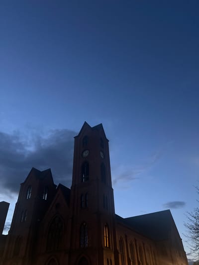 The image shows a brick church building against a blue sky. The church has a tall tower with clock faces visible on its sides. There are arched windows and other architectural details on the building. Some windows are illuminated from within. The sky is a gradient of blue, with some clouds present. The photograph appears to be taken from a low angle, looking up at the church. A few bare tree branches are visible in the upper right corner. The church is the Dutch Reformed Church, located in the city of Newburgh, New York.