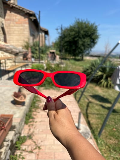 A person's hand, with dark red nail polish, holds a pair of red-framed sunglasses with black lenses in front of a blurred outdoor scene. The background features a brick building, green trees, and a bright blue sky. A pathway made of red bricks and some stone walls are also visible. The image appears to be taken during the daytime, with sunlight casting shadows. The location is specified as Cannara, Italy.