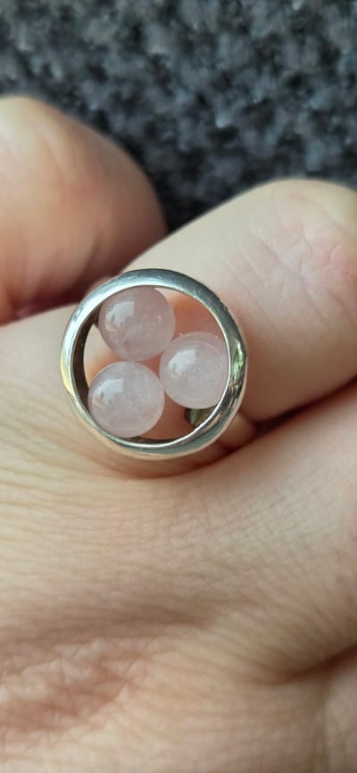 A close-up, high-angle shot shows a person's hand wearing a ring. The ring is made of silver and has a circular opening containing three small, smooth, pinkish-white stones. The background consists of a dark, textured surface. The image appears to be taken indoors, with the lighting casting soft shadows. There is no indication of the location being in Kehl, Germany.