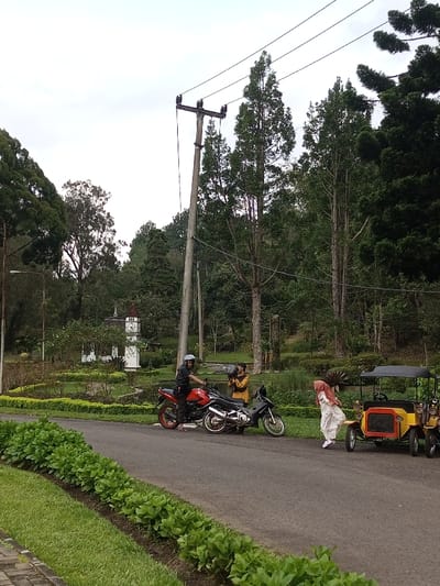The image depicts an outdoor scene in Ambositra, Madagascar, under an overcast sky. A paved road curves through the midground, bordered by a strip of green grass and low shrubs on the left.

On the road, in the midground, a male figure wearing a light-colored helmet, black jacket, and dark pants sits astride a red motorcycle, facing right. To his right, a second dark-colored motorcycle is parked. Behind the red motorcycle, a male individual in a yellow long-sleeved shirt stands, holding and operating a video camera pointed towards the right. Further right on the road, a female wearing a light-colored patterned dress and a pink hijab stands next to a yellow and black antique-style pedal car with a black canopy; her left leg is bent, indicating an action of stepping or engaging with the vehicle.

To the left of the road, a landscaped garden area is visible, featuring manicured shrubs, a small body of water, and a prominent white, miniature clock tower structure with a brown roof and visible clock faces. A street light pole stands near the garden's edge. The background is dominated by dense, tall green trees, suggesting a heavily wooded or forested area. A utility pole with multiple power lines is centrally positioned in the upper midground against the sky and tree line.
