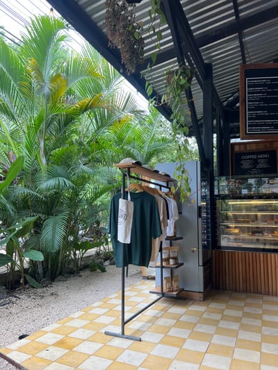 The image shows a clothing rack with three shirts hanging on it, two of them are green and one is white. The clothing rack is placed against a tiled wall with black and white square tiles in the outdoors. The scene appears to be located in front of a cafe, with a cafe menu and a glass display case filled with baked goods behind the clothing rack. There are plants in pots hanging from the ceiling, and a large tropical palm tree visible on the left side of the frame. The cafe is located in Santa Fe, Costa Rica, with the latitude: 9.642 and longitude: -85.165. There are no people visible in the scene. The image appears to be taken from a street view angle. The time of day appears to be daytime, with the sun shining brightly.