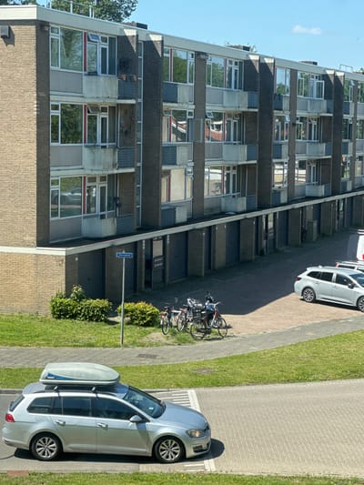 An eye-level, slightly angled, color photograph depicts a street scene in Breda, Netherlands, dominated by a multi-story residential building. The building, constructed of brick and concrete, features a repetitive pattern of recessed balconies and large windows. The windows reflect the surrounding environment, including the sky and trees. A street sign reading "Hoogeind" is visible near the base of the building.
To the right of the building, a narrow paved path runs parallel. Two cars are parked along the path, one silver and one white. Three bicycles are parked near the base of the building, close to the street sign. A strip of grass borders the path, with small bushes and trees providing greenery. The overall light suggests it is daytime.