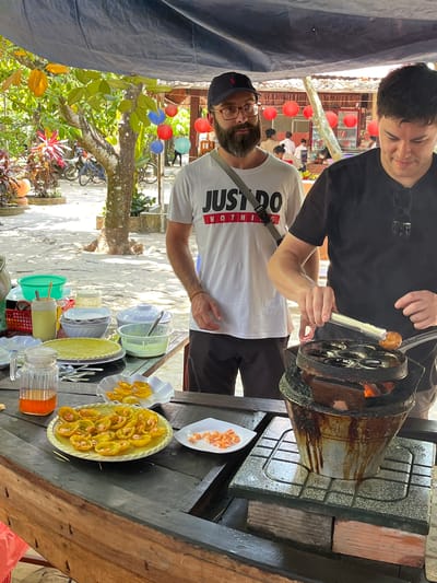Two men are cooking a snack dish in Tân Thạch, Vietnam. One man, wearing a black shirt and sunglasses, is using tongs to remove a small, round snack from a multi-hole cooking apparatus. Next to him, another man in a white t-shirt with "JUST DO NOTHING" printed on it, is assisting. Several plates of cooked snacks, and ingredients are laid out on a wooden table in front of them. The scene is located outdoors, with trees and other decorations visible in the background.