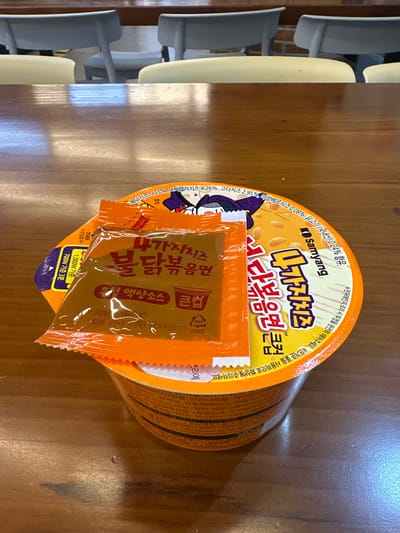 In the city of Jeonju-si, South Korea, an overhead shot showcases a wooden table with a cup of instant noodles. The cup has a bright orange lid and a label featuring Korean text and imagery of a cartoon character with a purple hat. Two smaller orange packets are placed on top of the cup lid. One packet is predominantly orange with Korean text and an image of a cup. The other packet is partially visible behind the first and appears to be a sauce packet. The background shows blurred light-colored chairs, suggesting an indoor setting, possibly a cafe or restaurant.