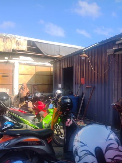A low-angle medium shot captures an outdoor scene with parked motorcycles in the foreground and a rustic building complex in the background. The sky above is a clear blue with a few wispy white clouds.

In the foreground, multiple motorcycles are densely parked. The closest motorcycle, viewed from the rear, is orange and grey with "FINO Sporty" branding. Partially visible is a motorcycle with a black helmet and "YAMAHA" branding. To its left, a motorcycle with a red seat and "HONDA" branding is present. A bright green motorcycle is also visible. The seats of these motorcycles are mostly black.

In the mid-ground, a woman wearing a brown hijab and dark clothing stands near an open doorway. She is looking to her right and holding a smartphone in her right hand. Behind her, the building appears to have large, light-colored doors or panels.

The background consists of two adjoining buildings. The building on the left has a white-colored facade with large, light-colored doors. The building on the right is constructed of corrugated metal, painted in a reddish-brown, weathered hue. A hanging lamp with an orange shade is attached to the corrugated metal wall, its cord trailing downwards. Two wooden poles lean against the corrugated metal wall, and an orange rectangular object is mounted on the wall.

The ground appears to be unpaved and uneven, with some debris scattered around. The overall impression is of a functional, possibly commercial or residential area in a warm climate. The scene is likely located in the city of Pompanua, Indonesia, given the context.