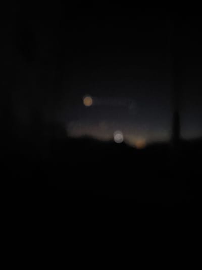 The image captures a blurred, dark scene with two circular, out-of-focus lights in the sky. A silhouetted structure, possibly a tower or building, is faintly visible on the right side of the horizon. The foreground is completely black and devoid of detail. The overall impression is that of a nighttime or very low-light photograph taken in a city, with the bokeh effect suggesting a shallow depth of field or a focus on distant lights. The specific location in Areni, Armenia cannot be determined from the visual information provided.