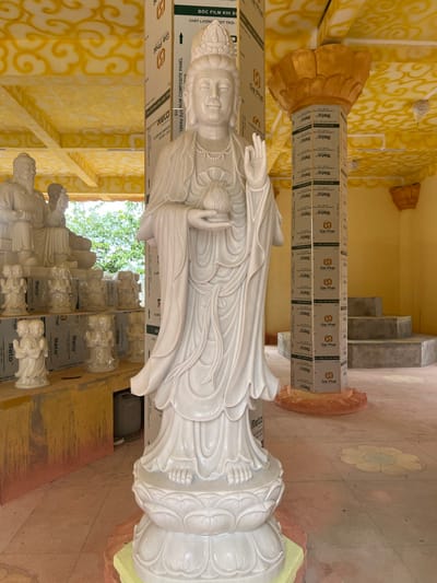 A large white marble statue of a standing female deity, likely Quan Âm (Guanyin), occupies the foreground of an indoor or semi-enclosed space in Đăk Mar, Vietnam. The statue features a crown, flowing robes, a beaded necklace, and holds a lotus bud in its left hand while its right hand is raised in a mudra. It rests on a multi-tiered lotus pedestal.
To the left of the main statue, a collection of smaller white marble or stone statues are arranged on shelves and directly on the floor. These include seated figures, some appearing to be wrapped in protective plastic, and several pairs of standing figures.
The environment exhibits characteristics of a construction or renovation site. Several structural pillars are prominently visible, wrapped in protective white panels branded with "Gia Phát" and "ALUMINUM COMPOSITE PANEL." The ceiling is painted a bright yellow with intricate, swirling patterns, and a large golden lotus-shaped capital adorns one pillar. The floor is a light-colored, possibly concrete, surface with faded circular designs. In the background right, a set of unadorned concrete steps leads upwards. Green foliage is visible through an opening on the left, indicating an external view.