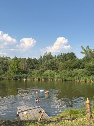 In Kamianka-Buzka, Ukraine, on a sunny day, several people are enjoying a swim in a calm body of water. The water is bordered by lush green trees and reeds. In the foreground, a wooden dock lies on the grassy bank, with several pairs of flip-flops scattered on its surface. In the water, two adults are visible, one appears to be holding a child. Two inflatable rings, one orange and one white, are also floating in the water, suggesting recreational activity. The sky above is a clear blue, dotted with fluffy white clouds.