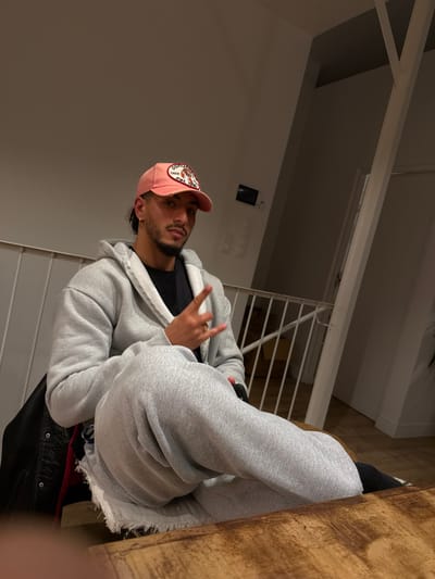 A male individual is seated indoors, observed from the waist up. He wears a pink baseball cap featuring an oval patch with a tiger graphic and partially legible text, a black t-shirt, and a light grey zip-up hoodie. Light grey sweatpants with a raw or distressed hem are visible on his lap, and black socks are present on his feet. A dark jacket is draped behind him. He has dark, short hair, a neatly trimmed beard and mustache, and a gold-colored ring on the ring finger of his right hand. His right hand is raised, forming a two-finger "peace" gesture, and he is looking directly at the camera. He is positioned behind a light brown wooden table, which occupies the lower foreground of the image. The background consists of light-colored walls. A white metal railing or balustrade is visible behind him, suggesting a multi-level interior space. A diagonal white structural beam or column extends from the right of the frame towards the top, and an electrical fixture is visible on the wall in the upper right. The lighting is artificial, illuminating the scene. The specific location within the city of Saint-Denis, France, cannot be determined from the visual information provided, as the image depicts a generic indoor residential or commercial setting.