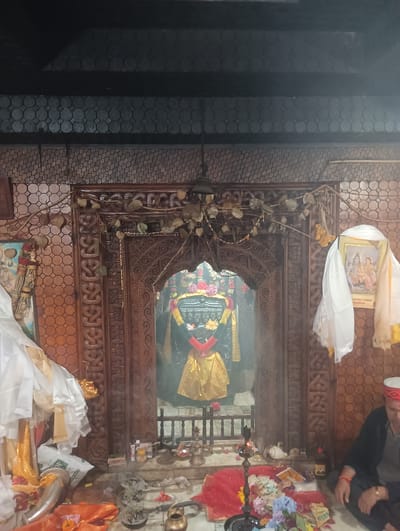 The image depicts the interior of a shrine, likely a Hindu temple, given the religious iconography present. At the center, behind a intricately carved wooden doorway, is a dark, upright stone idol, possibly a deity or lingam, adorned with a golden garment and red and green fabric. The shrine is decorated with garlands of dried leaves and yellow flowers. To the left of the doorway, a framed picture of a deity is visible, draped with white and yellow cloth. On the right side of the shrine, another framed picture of deities is hung, also draped with white and yellow cloth. In the foreground, offerings are laid out on the floor, including money, flowers, and other items commonly used in religious rituals. A person, appearing to be a male, is seated on the floor to the right of the shrine, dressed in dark clothing and wearing a traditional cap. The environment appears to be indoors, with patterned walls and a dark ceiling. The overall atmosphere suggests a place of worship, with evidence of ongoing religious practices. The image does not contain any visual cues that specifically identify the location as Gojra, India.